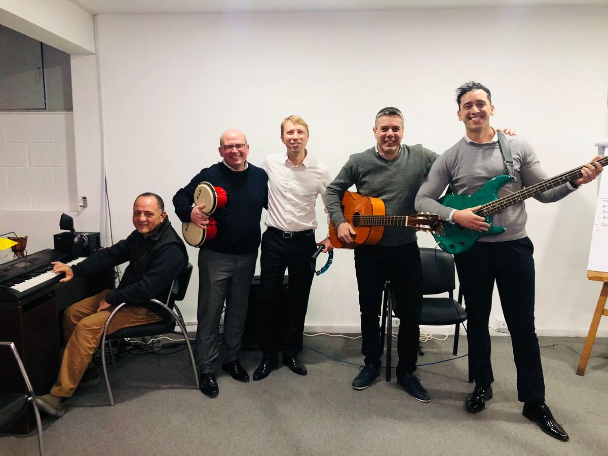 Cinco personas de oficina en una sala de reunión, con guitarra criolla, bajo, pandereta y teclado.
