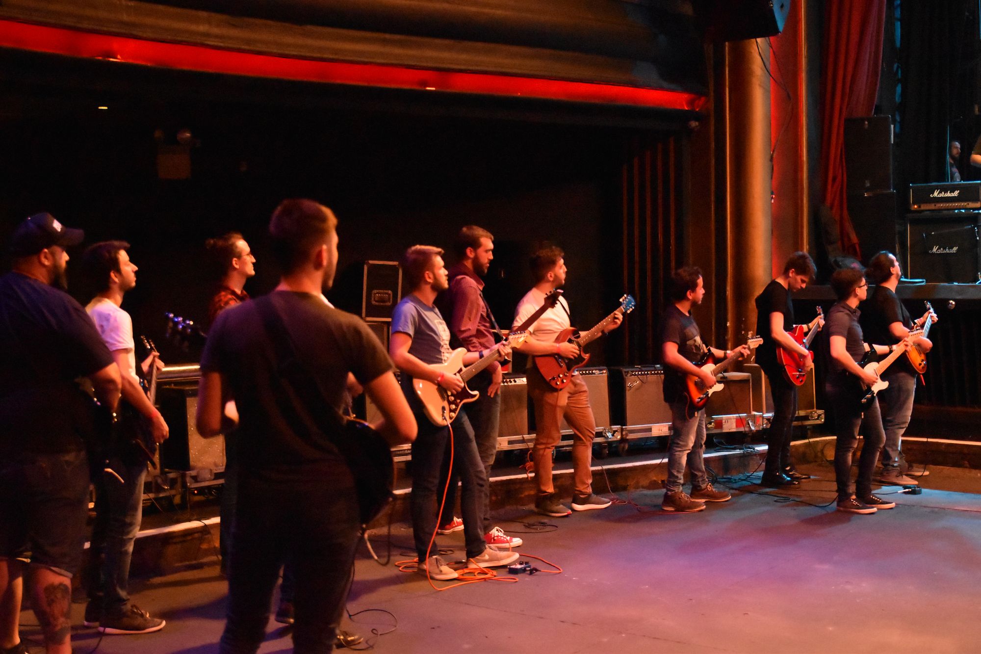 Doce personas de pie en el escenario del teatro Vorterix, cada una con una guitarra eléctrica, bajo luces rojas.